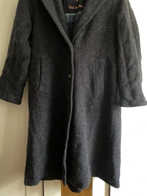 Mörkgrå ullkappa med huva - Stilren mörkgrå kappa från Ursel D. Atelier i ull med stor huva och knäppning framtill. Kappan har två fickor, dekorativt bälte bak och en rak, något loose passform. Perfekt för kyliga dagar och enkel att matcha med olika outfits.