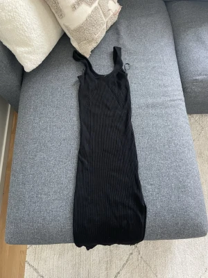 Svart ribbad ärmlös klänning - Säljer en svart, ribbad klänning med smala axelband och slits på sidan. Klänningen har en kroppsnära passform och är tillverkad i ett stretchigt material som ger en snygg siluett. Perfekt för dig som gillar minimalistisk stil.