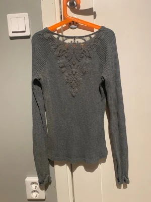 Grå ribbad tröja med spetsdetalj - Snygg grå långärmad tröja med ribbad struktur och unik spetsapplikation i blommönster över bröstet och axlarna. Tröjan har rund halsringning och smal passform, perfekt för dig som gillar detaljer och stilren design. Bra skick! 