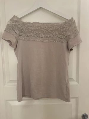 Beige offshoulder topp med spets - Söt beige offshoulder topp med bred spetsdetalj över axlarna. Toppen har korta ärmar och är tillverkad i mjukt material som känns skönt mot huden. 