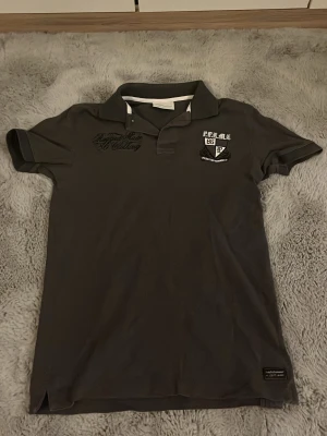 Mörkgrå pikétröja Peak Performance - Mörkgrå pikétröja från Peak Performance med klassisk krage och korta ärmar. Broderad logga och text på bröstet samt liten etikett nertill. Perfekt för en sportig och stilren look.