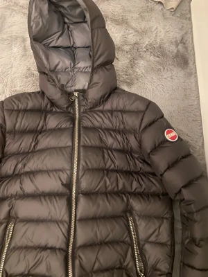 Svart dunjacka från Colmar - Snygg svart dunjacka från Colmar med huva och quiltad design. Jackan har dragkedja framtill och på fickorna samt en rund Colmar-logga på ärmen. Perfekt för kalla dagar och riktigt stilren look.