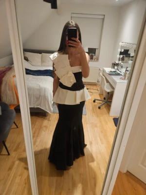 Svart och vit långklänning med volang - Säljer en unik långklänning i svart och vitt med stora volangdetaljer och en axelbandslös design. Klänningen har en markerad midja med svart dekoration och en dramatisk, asymmetrisk överdel. Perfekt för dig som vill sticka ut.