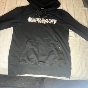 Svart Represent hoodie med blommor - Svart hoodie från Represent med stor blommig logga på bröstet och en stor bokstav R med blommor och fågel på ryggen. Tröjan har huva med snörning och en klassisk magficka. Perfekt för dig som gillar streetwear med lite extra detaljer.