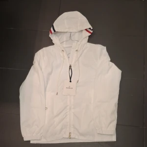Vit windbreaker från Moncler med huva - Snygg vit windbreaker från Moncler med huva och dragkedja framtill. Jackan har snörning vid huvan och Moncler-logga på ärmen. Insidan har en Moncler-etikett och detaljer i rött, vitt och blått på huvan. Perfekt för dig som gillar stilrena och sportiga plagg.