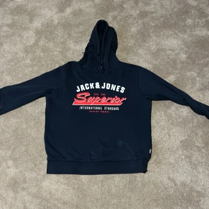 Mörkblå hoodie Jack & Jones Superior - Snygg mörkblå hoodie från Jack & Jones med trycket 'Superior' i rött och vitt på bröstet. Storlek M