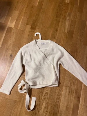 Vit omlottopp från NA-KD - Säljer en vit omlottopp från NA-KD i storlek M. Toppen har långa ärmar, v-ringning och knytband i midjan. Materialet är mjukt och lite fluffigt, vilket gör den extra mysig. Perfekt för dig som gillar stilrena och enkla plagg med en twist.