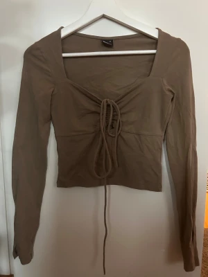 Beige/brun långärmad topp med snörning - Säljer en beige/brun långärmad topp med snygg snörning framtill och croppad passform. Toppen har en fyrkantig urringning och är gjord i mjuk bomull som känns skön mot huden. Perfekt för dig som gillar trendiga detaljer och neutral färg.