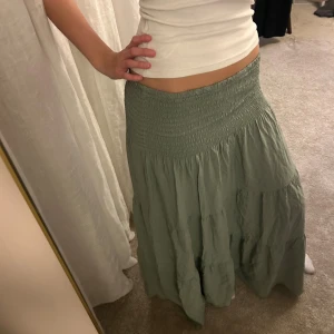 Grön långkjol med smock i midjan - Säljer en flowy grön långkjol med bred smock i midjan som ger en skön och flexibel passform. Kjolen har volanger och är tillverkad i ett mjukt, lätt tyg som passar perfekt till varma dagar. Enkel att matcha med crop tops eller linnen.