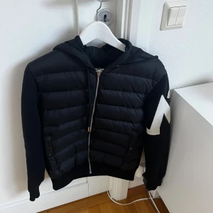 Svart Moncler cardigan med huva - En svart cardigan från Moncler med huva och dragkedja fram. Den har stickade ärmar med ett vitt M och Moncler-loggan på ena ärmen. Framsidan är dunfylld för extra värme, och den har två fickor med knappar. En snygg, sportig och riktigt användbar jacka för kyliga dagar. Båda  qr kod o nfc funkar
