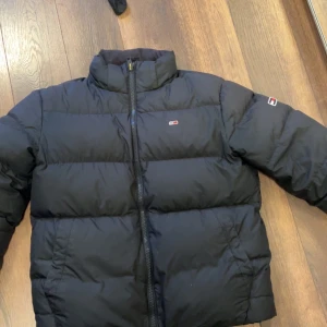 Svart pufferjacka från Tommy Jeans - Svart pufferjacka från Tommy Jeans med hög krage och diskret logga på bröstet och ärmen. Jackan har dragkedja framtill och är quiltad för extra värme. Perfekt för kalla dagar och har en clean, stilren look som funkar till jeans eller cargo.