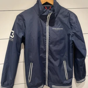 Blå vindjacka från Race Marine - Snygg marinblå vindjacka från Race Marine med vita detaljer och dragkedjor. Jackan har hög krage, två fickor med dragkedja och logga på bröstet samt ärmen. Perfekt för blåsiga dagar och sportiga outfits.