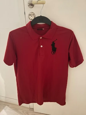 Röd Polo pikétröja med svart logga - Snygg röd pikétröja från Polo i 100% bomull. Klassisk krage, två vita knappar och stor svart broderad logga på bröstet. Kortärmad och perfekt för en chill och stilren look. Passar dig som gillar sportig vibe och enkel design.