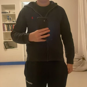 Mörkblå hoodie från Polo Ralph Lauren - Storlek 10/12 Snygg mörkblå hoodie från Polo Ralph Lauren med röd broderad logga på bröstet. Tröjan har huva, dragkedja framtill och två fickor. Tillverkad i mjuk bomullsmix och har långa ärmar med ribbade muddar. Perfekt för chill dagar och streetstyle.