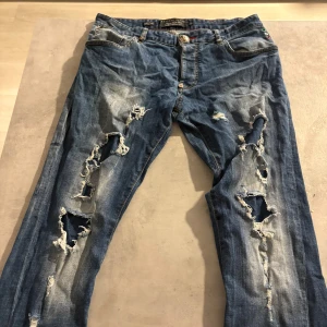 Philipp Plein blå ripped jeans straight - Blå jeans från Philipp Plein med super straight cut och coola slitningar framtill. Snygga detaljer som röd söm, metallknapp med fågelmotiv och bakficka med läderpatch och korslagda ben. Riktigt edgy streetstil för dig som gillar statement-plagg.