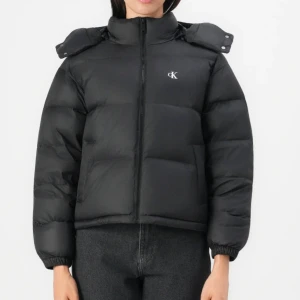 Svart pufferjacka från Calvin Klein - Snygg svart pufferjacka från Calvin Klein med hög krage, avtagbar huva och diskret CK-logo på bröstet. Jackan har dragkedja framtill och elastiska muddar vid ärmsluten. Perfekt för kalla dagar och ger en cool, modern look. Köpte den för runt 2700 och säljer den för 2000 men pris kan diskuteras. Använd fåtal gånger så gott som ny.