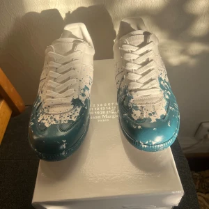 Maison Margiela sneakers med blå färgstänk - Tja! Dem enda sällsynta Maison Margiela skorna som finns i plick! och inte säljs längre på nätet, i vitt skinn med unika blå färgstänk över tån och sidorna. Snygg och konstnärlig vibe som sticker ut, perfekt för dig som gillar statement pieces!