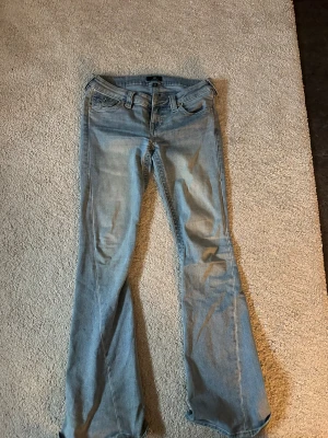 True Religion Joey Low Rise Flare Jeans, W26 - Snygga ljusblå jeans från True Religion, modell Joey Low Rise Flare. De har låg midja, klassisk femficksdesign och utställda ben. Ikoniska bakfickor med broderad logga och silverfärgade knappar. De har dessvärre små slitningar i slutet av benen (se bild 5), men det är inget som förstör helhetsbilden. Nypris 1300kr