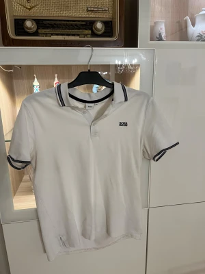 Vit pikétröja från Hugo Boss - Vit pikétröja från Hugo Boss med klassisk krage och svarta detaljer på krage och ärmslut. Tröjan har korta ärmar, knäppning framtill och diskret Boss-logga på bröstet. Tillverkad i bomull för en skön och stilren känsla.( Den är liten i storleken)