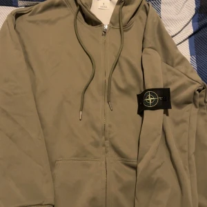 Beige hoodiejacka från Stone Island - Säljer en beige hoodiejacka från Stone Island med dragkedja framtill och klassisk logotyp-patch på ärmen. Jackan har huva med snörning och känguruficka. Perfekt för dig som gillar streetwear och vill ha en clean look. Storlek M, priset kan diskuteras.