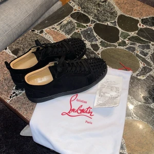 Christian Louboutin svarta sneakers med nitar - Svarta sneakers från Christian Louboutin i mocka med coola svarta nitar på tån och klassisk röd sula. Skorna har snörning och rund tå, samt en platt sula. Perfekt för dig som vill sticka ut med exklusiv design och edgy detaljer.