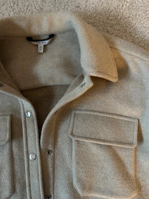 Beige jacka från & Other Stories, 34 - Säljer en beige jacka från & Other Stories i storlek 34. Jackan har två stora bröstfickor med lock, klassisk krage och tryckknappar framtill. Materialet är mjukt och känns ulligt, perfekt för lager på lager.