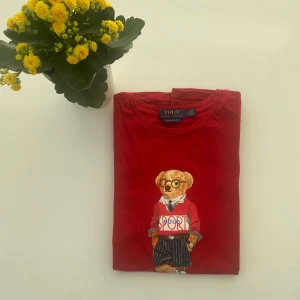 Röd t-shirt med björntryck från Polo Ralph Lauren - Säljer en röd t-shirt från Polo Ralph Lauren med ett stort tryck av en björn i sportig outfit på framsidan. T-shirten har rund halsringning och är i modellen custom slim fit. Perfekt för dig som gillar streetstyle och klassiska märken.