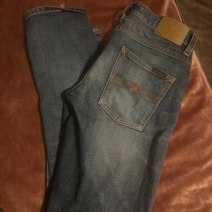Blå jeans från Nudie Jeans - Snygga blå jeans från Nudie Jeans med klassisk femficksdesign och coola slitningar framtill och baktill. Jeansen har orangea sömmar och Nudie-logga på bakfickan. Passformen är rak och materialet är mjukt denim i bomull.