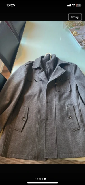 Grå rock med stickad krage - Snygg grå rock med klassisk design och stickad krage. Jackan har både knappar och dragkedja framtill, samt flera fickor med knappstängning. Materialet är ullblandning och insidan har en mjuk stickad del som ger extra värme. Perfekt för kyliga dagar. Kragen är avtagbar.  Jackan är helt nyoch kostar 999kr nypris. 