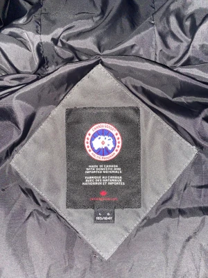 Svart/Grå pufferjacka från Canada Goose - Säljer en svart/grå  pufferjacka från Canada Goose med huva och klassisk logga på ärmen. Jackan har dragkedja framtill, stora fickor och quiltad design. Perfekt för kalla dagar och riktigt snygg streetstil.