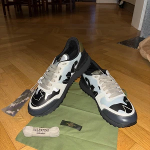 Valentino Rockrunner sneakers camo - Snygga Valentino Rockrunner sneakers med svart, grå och vit camouflage-mönster. Skorna har rund tå, platt sula med mönstrad undersida och beige snörning. Mix av skinn och mocka med gulddetalj på hälen. Perfekt för dig som vill sticka ut med exklusiva sneakers.