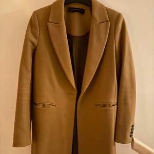 Beige kappa från Zara Woman - Stilren beige kappa från Zara Woman med klassisk krage och knäppning framtill. Jackan har två fickor och dekorativa knappar vid ärmsluten. Perfekt för dig som gillar en clean och tidlös look.