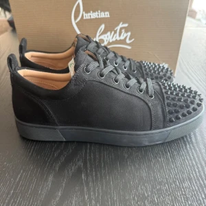 Christian Louboutin svarta sneakers med nitar - Svarta sneakers från Christian Louboutin i mocka med rund tå och snörning. Tåpartiet är täckt av svarta nitar för en edgy look. Sulan är svart med den ikoniska röda undersidan och Louboutin-logga. Insidan är ljusbeige och skon har en klassisk låg profil.