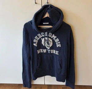 Marinblå hoodie Abercrombie & Fitch - Snygg marinblå hoodie från Abercrombie & Fitch med broderad tiger och texten 'Abercrombie New York' på bröstet. Tröjan har huva med snörning, känguruficka och långa ärmar. Perfekt för dig som gillar streetwear och vill ha en chill look.