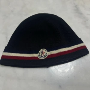 Svart Moncler mössa med ränder - Snygg svart  mössa från Moncler med klassiska röda, vita och blå ränder längs kanten. Mössan har Moncler-logga framtill och är stickad i ett mjukt material som håller dig varm och stilren under kalla dagar.