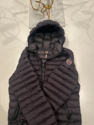 Svart pufferjacka från Colmar - Snygg svart pufferjacka från Colmar med huva och dragkedja. Jackan har quiltad design, två sidofickor med dragkedja och Colmar-logga på ärmen. Perfekt för kalla dagar och riktigt skön att bära. Materialet är lätt och smidigt syntet. Bara använts fåtal gånger 