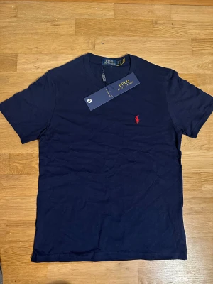 Marinblå t-shirt från Polo Ralph Lauren - Snygg marinblå t-shirt från Polo Ralph Lauren i mjuk bomull. Klassisk passform med korta ärmar och rund hals. Ikoniska röda broderade polospelaren på bröstet ger en stilren touch. Perfekt för en clean och tidlös look.