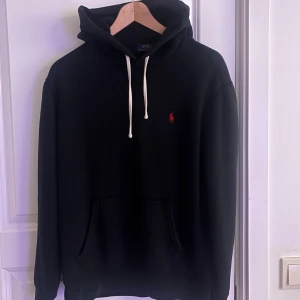 Svart hoodie från Polo Ralph Lauren - Svart hoodie från Polo Ralph Lauren med klassisk röd logga broderad på bröstet. Tröjan har vit snörning i huvan, känguruficka och långa ärmar. Perfekt för en chill och stilren look.