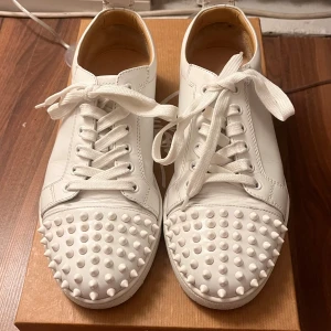 Christian Louboutin vita sneakers med nitar - Säljer ett par vita sneakers från Christian Louboutin med ikoniska röda sulor och coola nitar på tårna. Skorna har snörning och är tillverkade i skinn med platt sula och rund tå. Perfekt för dig som vill sticka ut med en lyxig och edgy stil. Bought from Vestiare!