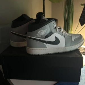 Nike Air Jordan 1 Mid grå/vit - Nike Air Jordan 1 Mid sneakers i stilren grå och vit färg med svarta detaljer. Klassisk Jordan-logga på sidan, perforerad tå och vit snörning. Skorna har en rund tå och platt sula, tillverkade i skinn och syntet. Perfekt för dig som gillar streetwear och snygg design. Är storlek 43 men sitter som 42. Låda följer även med.