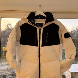 Vit och svart Sail Racing dunjacka - Säljer en fet dunjacka från Sail Racing i vit och svart med huva och coola detaljer. Jackan har två bröstfickor med dragkedja, broderad logga på huvan och patch på ärmen. Perfekt för kalla dagar och har en riktigt clean look. Jackan kommer även att kemtvättas vid köp!🧼