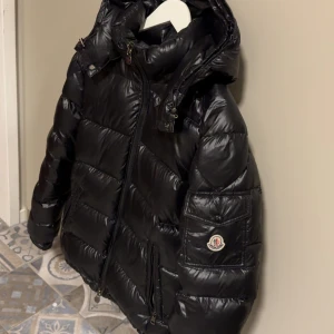 Svart Moncler pufferjacka med huva - Svart pufferjacka från Moncler med glansig finish, huva och dragkedja framtill. Jackan har Moncler-logga på ärmen, praktiska fickor med dragkedja och quiltad design. Perfekt för kalla dagar och har en cool, urban vibe.   Säljs pga att har tröttnat på den. Har tyvärr inget kvitto. Priset är lågt för att en lapp på insidan är halvt trasig.