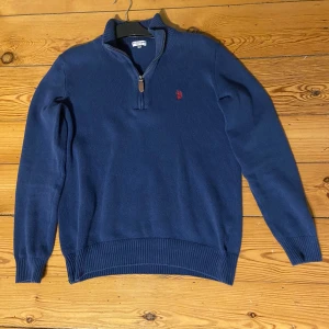Quarter zip  - Säljer en marinblå stickad tröja från U.S. Polo Assn med halv dragkedja och röd broderad logga på bröstet. Tröjan har ribbade muddar och hög krage, perfekt för kyliga dagar. Klassisk och stilren modell som funkar till det mesta.