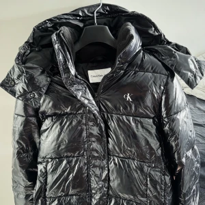 Svart pufferjacka från Calvin Klein - Snygg svart pufferjacka från Calvin Klein med glansig finish och broderad CK-logga på bröstet. Jackan har huva, dragkedja framtill och quiltad design som ger en modern vibe. Perfekt för kalla dagar när du vill hålla stilen.