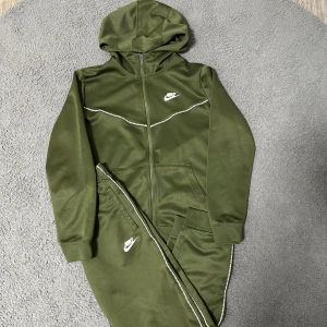 Grön set från Nike  - Snygg grön hoodie från Nike med vit logga på bröstet och matchande detaljer längs dragkedjan. Tröjan har huva, långa ärmar och praktiska fickor framtill. Materialet är mjukt och känns sportigt, perfekt för chill eller träning. Samma sak med byxorna, fast det finns en liten röd fläck på byxan som du kan se på sista bilden. Syns inte så mycket? 