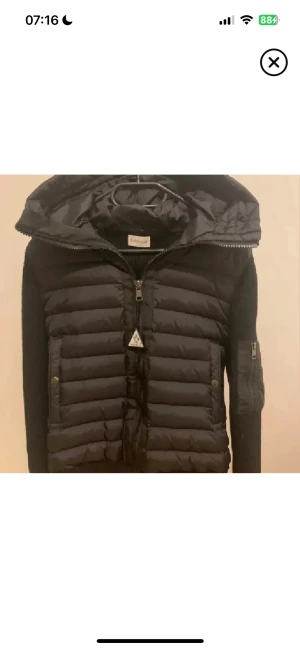 Svart Moncler pufferjacka med huva - Svart pufferjacka från Moncler med stickade ärmar och quiltad front. Jackan har en stor, vadderad huva och dragkedja framtill med Moncler-logga. Snygg kombination av syntet och stickat material, perfekt för höst och vinter. Fickor med dragkedja på ärmen och sidan.