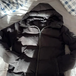 Svart pufferjacka Supply & Demand S - Svart pufferjacka från Supply & Demand med huva och snyggt emblem på ärmen. Jackan har dragkedja framtill och är vadderad för extra värme. Perfekt för kalla dagar och har en clean, urban stil. Passar dig som gillar streetwear och vill ha en jacka med attityd.