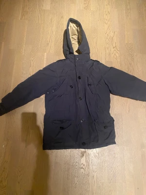 Svart parkajacka från Polo Ralph Lauren - Svart parkajacka från Polo Ralph Lauren med huva och flera fickor med knappar. Jackan är fodrad med beige insida och har en klassisk, rak passform. Materialet är en mix av bomull och nylon, med dunfyllning för extra värme. Perfekt för kalla dagar.