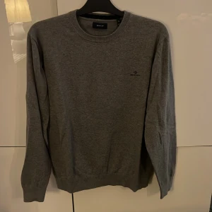 Grå stickad tröja från GANT XL - Snygg grå stickad tröja från GANT i storlek XL. Tröjan har rund halsringning, diskret logga broderad på bröstet och ribbade muddar vid ärmslut och nederkant. Perfekt för dig som gillar klassisk och enkel stil.