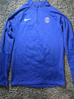Blå PSG långärmad Nike fotbollströja - Slim fit blå fotbollströja från Nike med PSG-logga på bröstet och vit swoosh. Tröjan har halv dragkedja fram och är tillverkad i Dri-Fit material som andas. Ärmarna har diskret mönster och tröjan är perfekt för träning eller match.